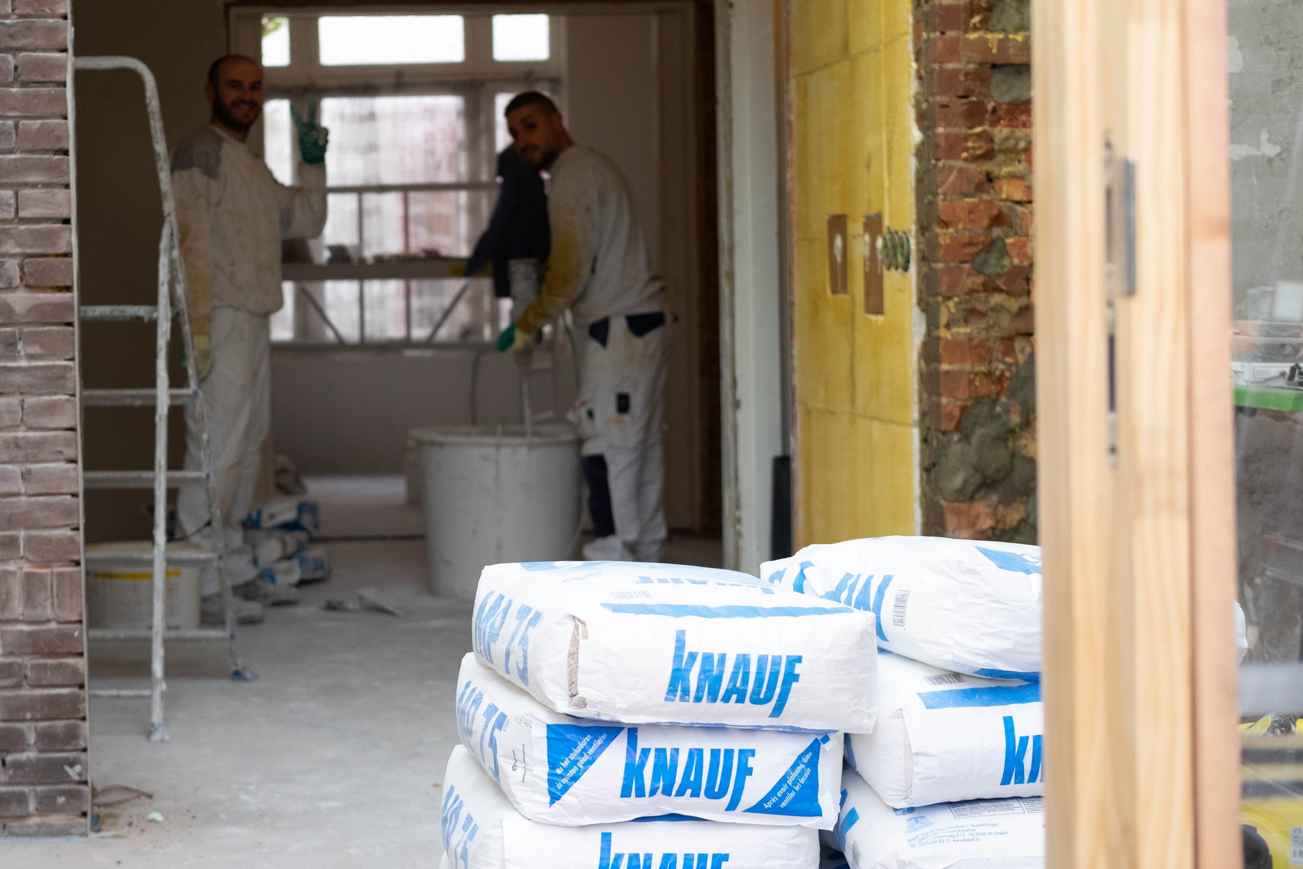 Murat en Kadir aan het werk in een kale ruimte, zakken Knauf stucmortel op de voorgrond